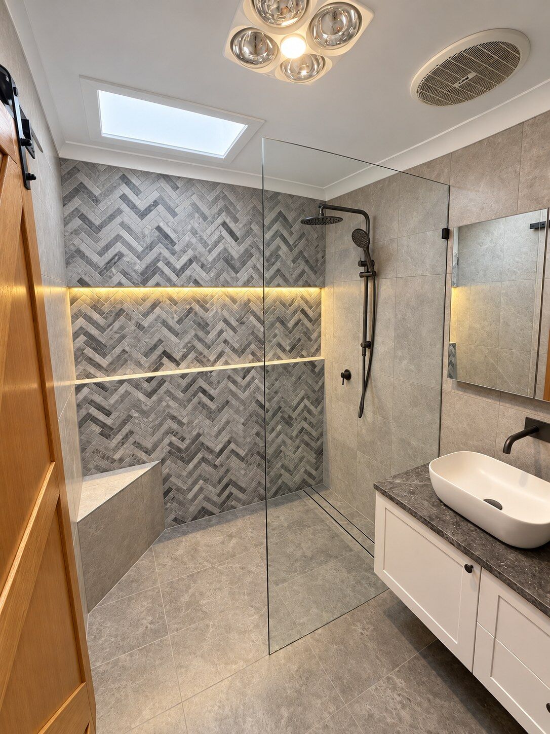 A bathroom with a herringbone tiled feature wall, a lit niche and a frameless shower screen